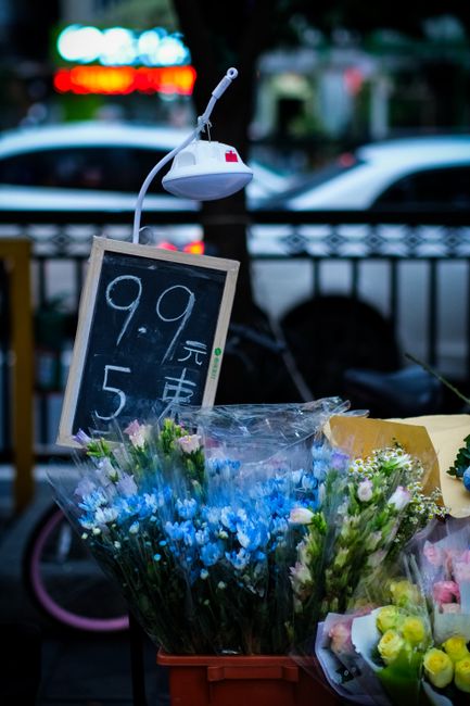 An outdoor display of fresh floral bouquets featuring a variety of flowers including blue, white, and pink blossoms arranged in clear plastic wrapping and placed in orange crates. The bouquets showcas