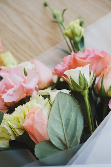 A close-up of a floral bouquet featuring pale pink and cream lisianthus flowers with soft, ruffled petals, complemented by fresh green foliage and greyish-green leaves. The bouquet is wrapped in clear