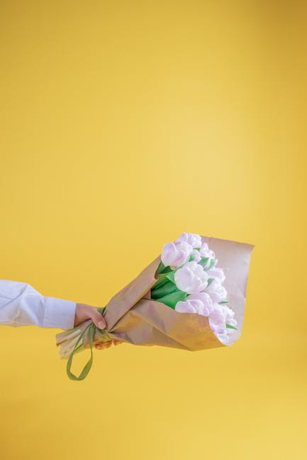 A hand holding a bouquet of fresh, pastel pink and white tulips wrapped in light brown craft paper with a green ribbon against a vibrant yellow background. The tulips have smooth, rounded petals and g