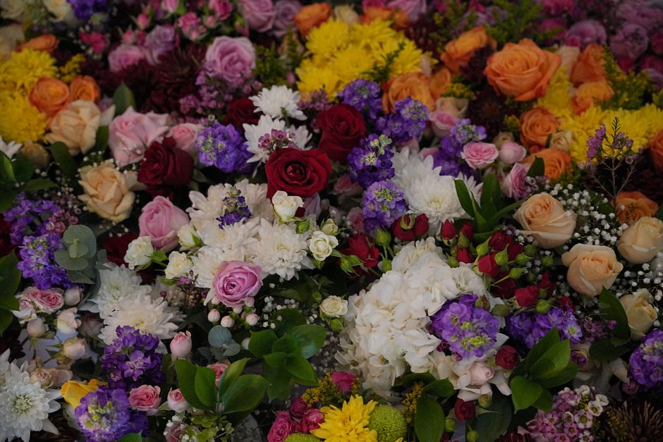 A vibrant assortment of fresh flowers arranged in a lush bouquet, featuring deep red roses, soft pink roses, white chrysanthemums, purple statice, yellow daisies, and sprigs of baby's breath, compleme
