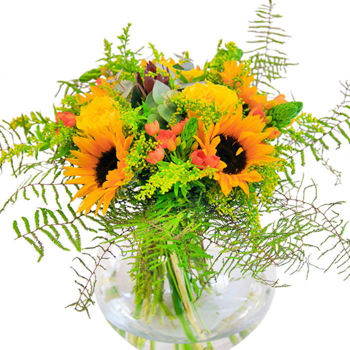 The instant impression of this glorious bouquet is pure sunshine, an explosion of yellow and orange hues reminiscent of Cockfosters gardens in peak bloom. Large, radiant sunflowers steal the eye first, their golden petals unfurling with cheerful confidence, while compact yellow garden roses nestle beside them, intricate spirals promising a timeless, welcoming beauty. The bouquet's deep green foliage and airy fern fronds create layers, with strands spilling lazily from the rim-much like the foliage one might find on gentle walks through nearby parks or the leafy corners of Bramley Road. Punctuating the vibrant yellow palette are glossy clusters of Hypericum berries-a splash of juicy red that enlivens the whole display. Delicate sprigs of pale green and soft blue-grey eucalyptus peek through, weaving rustic sophistication through the arrangement. Anchored in a crystal-clear glass vase that reveals the architecture of stems beneath the glistening surface, this composition is more than a simple arrangement-it's a declaration of happiness. The delicate scent of garden roses recalls Cockfosters' summer air, making this bouquet the perfect gift for anniversaries, housewarmings, or just to fill a home with warmth and vivacity.
